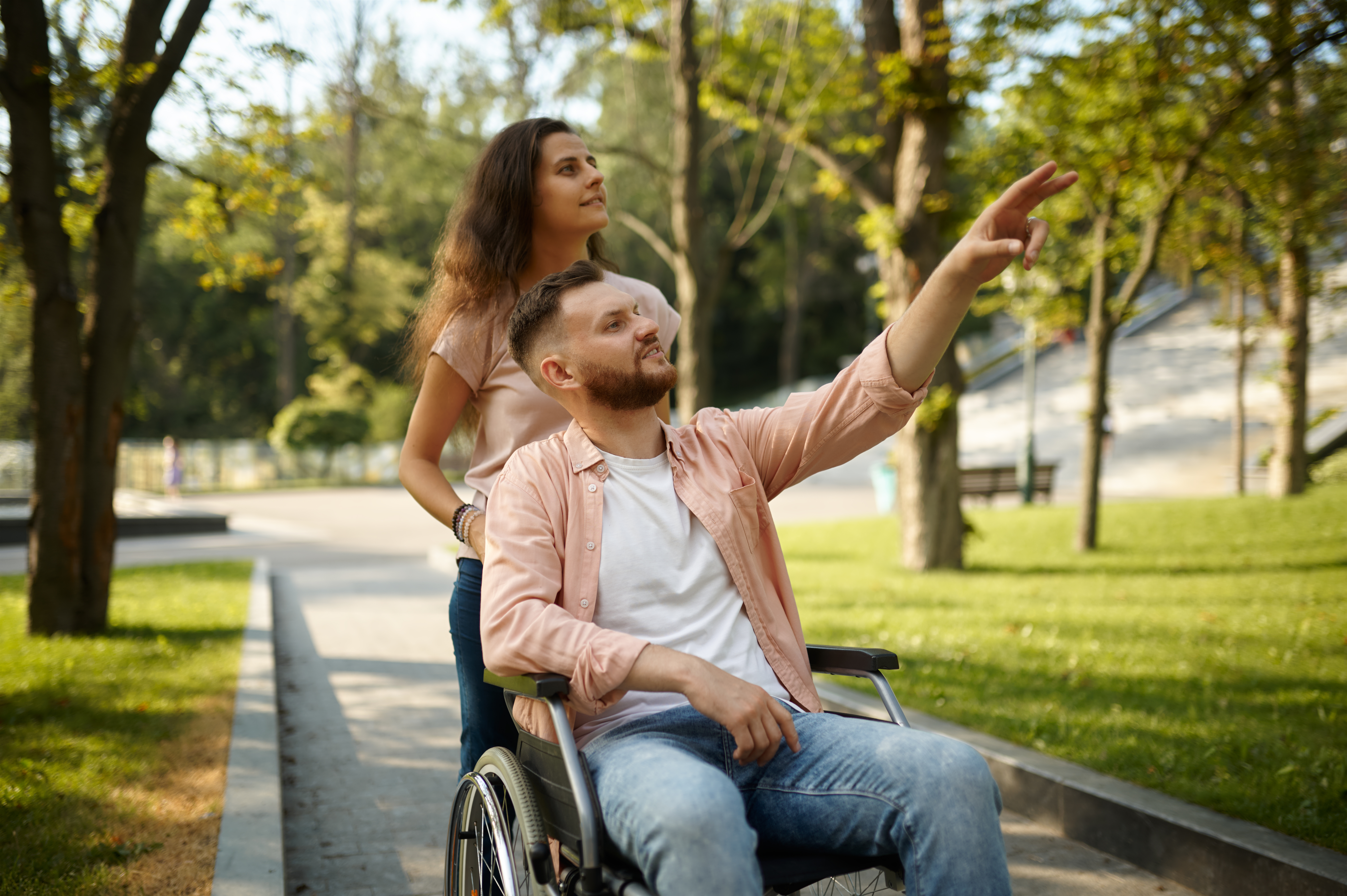 Young family couple with wheelchair walking in the park. Paralyzed people and disability, care for a disabled man. Husband and wife overcome difficulties together